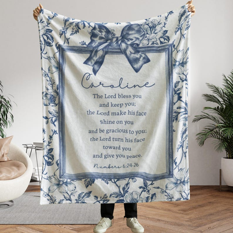 May include: A white and blue floral throw blanket with a bow design and the name "Caroline" in script. The blanket features a framed text with a religious quote from Numbers 6:24-26. The blanket is a soft, cozy home decor item.