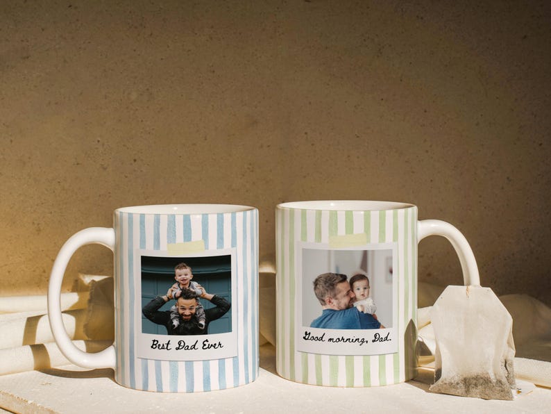May include: Two white ceramic mugs with striped patterns. One mug has blue stripes and the text "Best Dad Ever" with a photo. The other mug has green stripes and the text "Good morning, Dad" with a photo. A tea bag hangs from the second mug.