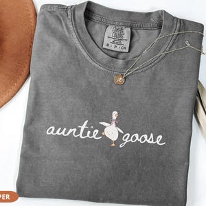 Puede incluir: Una camiseta gris con las palabras "auntie goose" en escritura blanca y una ilustraci&oacute;n de ganso de dibujos animados. Un collar dorado con un colgante descansa sobre la camiseta. La camiseta est&aacute; doblada sobre una superficie blanca.