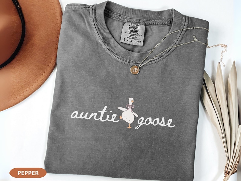 Puede incluir: Una camiseta gris con las palabras "auntie goose" en escritura blanca y una ilustraci&oacute;n de ganso de dibujos animados. Un collar dorado con un colgante descansa sobre la camiseta. La camiseta est&aacute; doblada sobre una superficie blanca.