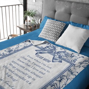 Pode incluir: Cobertor branco e azul floral com o texto "DEAR MOM" e uma mensagem sincera. O cobertor apresenta uma borda decorativa e um design de la&ccedil;o, perfeito para um quarto aconchegante.