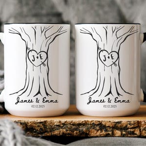 May include: Two white ceramic mugs with black handles and rims. Each mug features a black tree design with a heart containing the initials "J+E" pierced by an arrow. The names "James & Emma" and the date "02.12.2025" are printed below the tree.