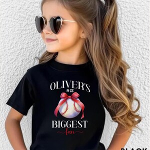 Puede incluir: Camiseta negra con el texto "OLIVER'S #22 BIGGEST fan" y un gr&aacute;fico de b&eacute;isbol con una cinta roja. La persona lleva gafas de sol en forma de coraz&oacute;n.