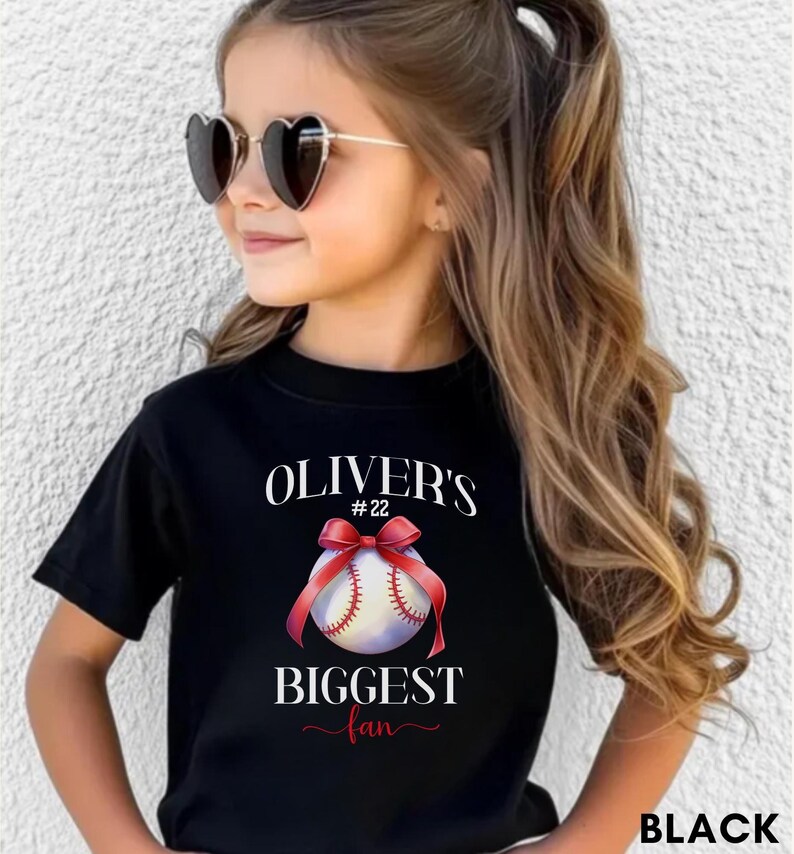 Puede incluir: Camiseta negra con el texto "OLIVER'S #22 BIGGEST fan" y un gr&aacute;fico de b&eacute;isbol con una cinta roja. La persona lleva gafas de sol en forma de coraz&oacute;n.