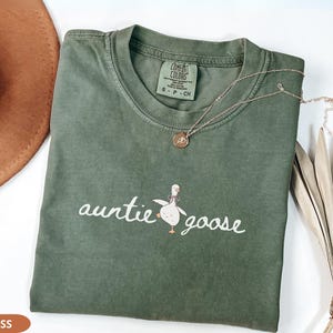 Puede incluir: Una camiseta verde musgo con las palabras "auntie goose" en cursiva, acompa&ntilde;ada de una ilustraci&oacute;n de un ganso de dibujos animados. Un collar de oro con un colgante descansa sobre la camiseta. Un sombrero marr&oacute;n es visible en el fondo.