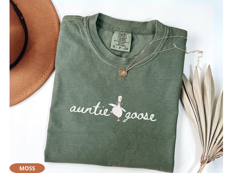 Puede incluir: Una camiseta verde musgo con las palabras "auntie goose" en cursiva, acompa&ntilde;ada de una ilustraci&oacute;n de un ganso de dibujos animados. Un collar de oro con un colgante descansa sobre la camiseta. Un sombrero marr&oacute;n es visible en el fondo.