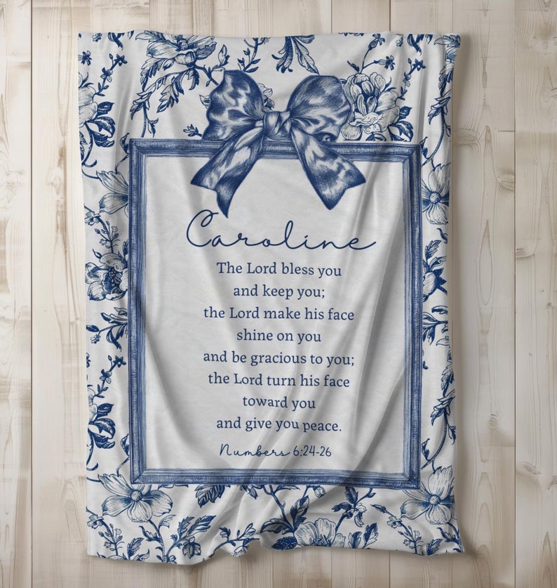 May include: A white blanket with a blue floral pattern and a blue bow. The blanket has a framed section with the name "Caroline" and a Bible verse. The verse is from Numbers 6:24-26.