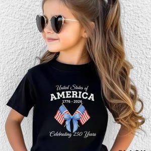 May include: Black t-shirt with the text "United States of AMERICA 1776-2026 Celebrating 250 Years" and a graphic of two American flags tied with a blue bow. The shirt is worn by a person wearing heart-shaped sunglasses.