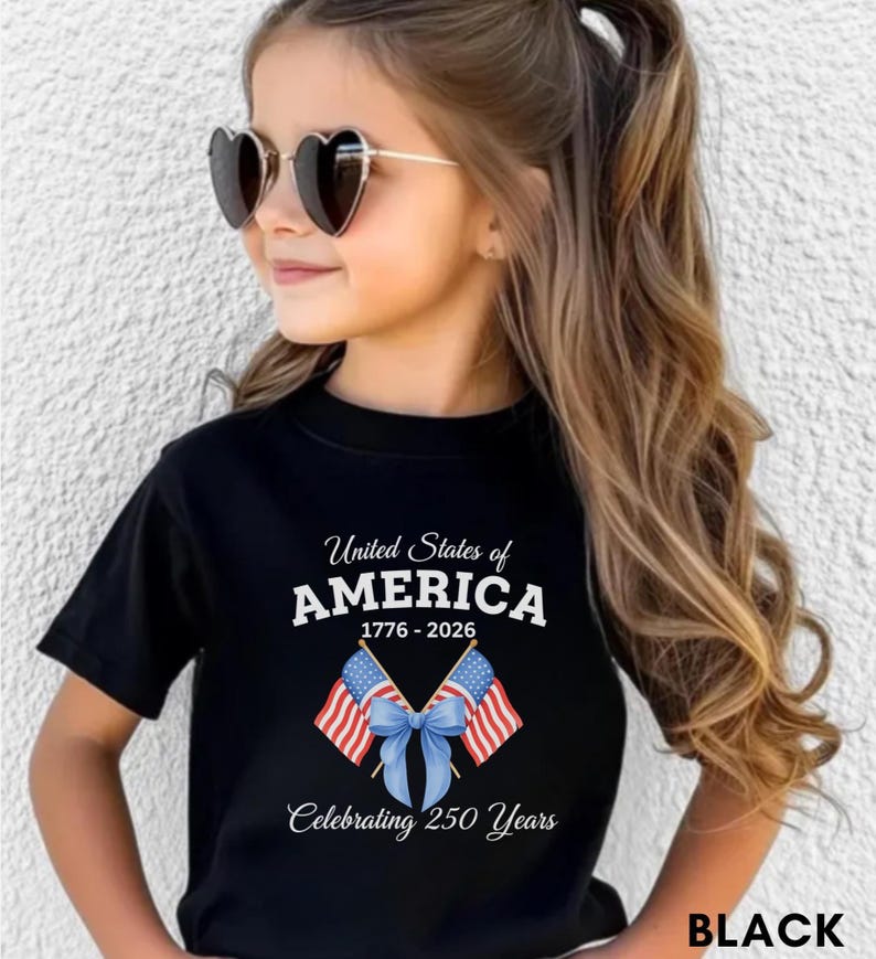 May include: Black t-shirt with the text "United States of AMERICA 1776-2026 Celebrating 250 Years" and a graphic of two American flags tied with a blue bow. The shirt is worn by a person wearing heart-shaped sunglasses.