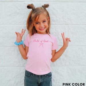 May include: A pink t-shirt with the text "Child of God" in a cursive font and a bow graphic. The shirt is paired with dark denim jeans. The image is set against a white brick wall.