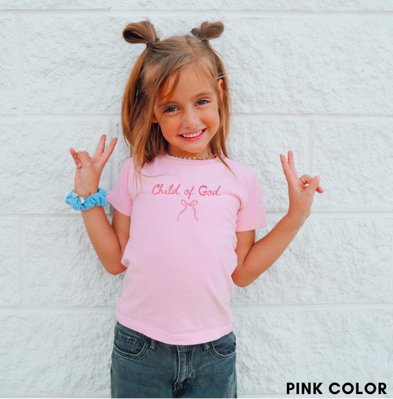 May include: A pink t-shirt with the text "Child of God" in a cursive font and a bow graphic. The shirt is paired with dark denim jeans. The image is set against a white brick wall.