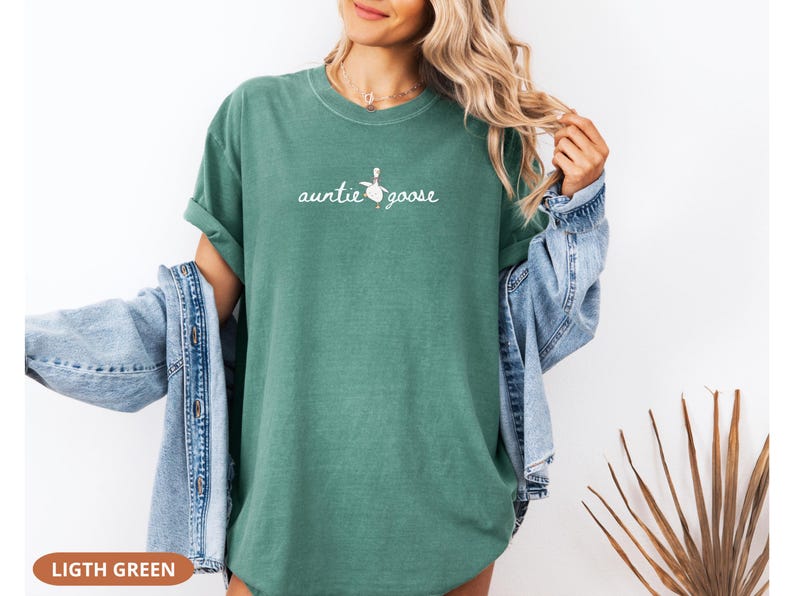 Puede incluir: Camiseta verde claro con la frase "auntie goose" en letra cursiva, acompa&ntilde;ada de una peque&ntilde;a ilustraci&oacute;n de un ganso. La camiseta es de color liso y tiene mangas cortas. El texto "LIGHT GREEN" est&aacute; en la parte inferior.