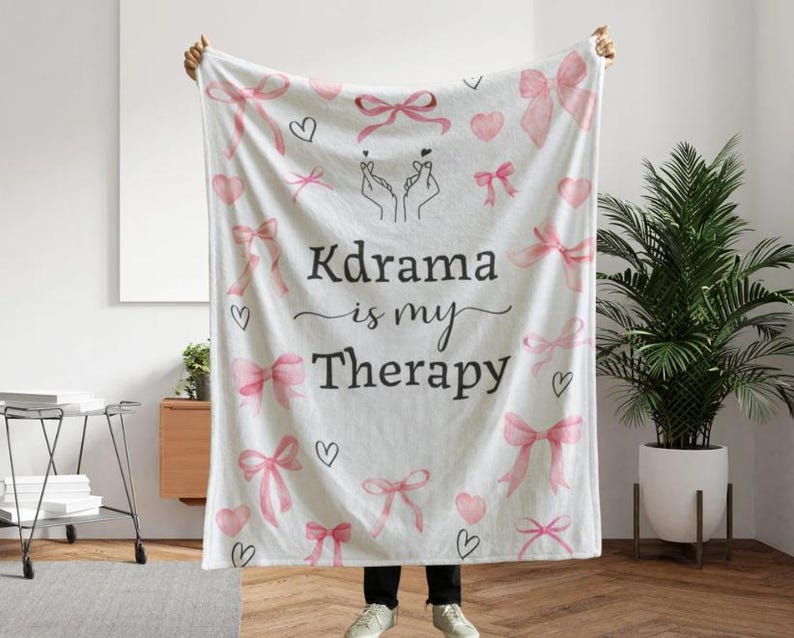 May include: White blanket with pink bows, hearts, and the text "Kdrama is my Therapy." The blanket features a hand-drawn design of two hands making a heart shape. A cozy and decorative item.