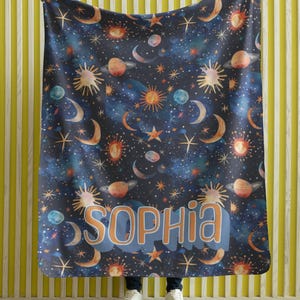 May include: A blue fleece blanket with a celestial pattern of stars, moons, planets, and suns. The blanket has the name "Sophia" printed in a blue and orange font.