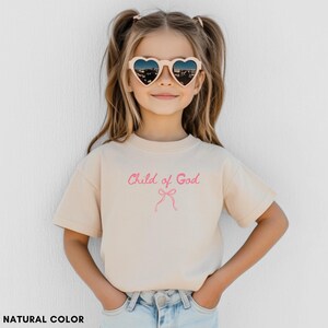 May include: A beige t-shirt with the text "Child of God" in pink script and a pink bow graphic. The shirt is worn by a young person wearing heart-shaped sunglasses.