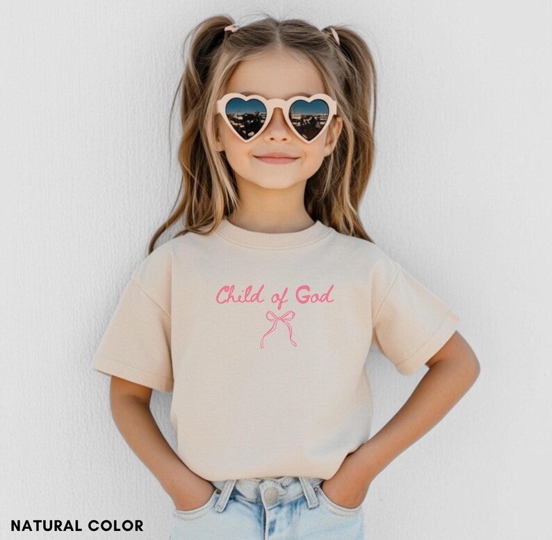 May include: A beige t-shirt with the text "Child of God" in pink script and a pink bow graphic. The shirt is worn by a young person wearing heart-shaped sunglasses.