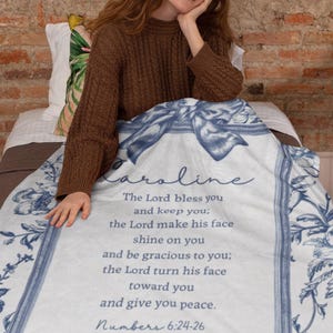 May include: A white and blue floral blanket with the name "Caroline" and a Bible verse printed on it. The verse reads "The Lord bless you and keep you... Numbers 6:24-26". The blanket has a decorative border and bow design.