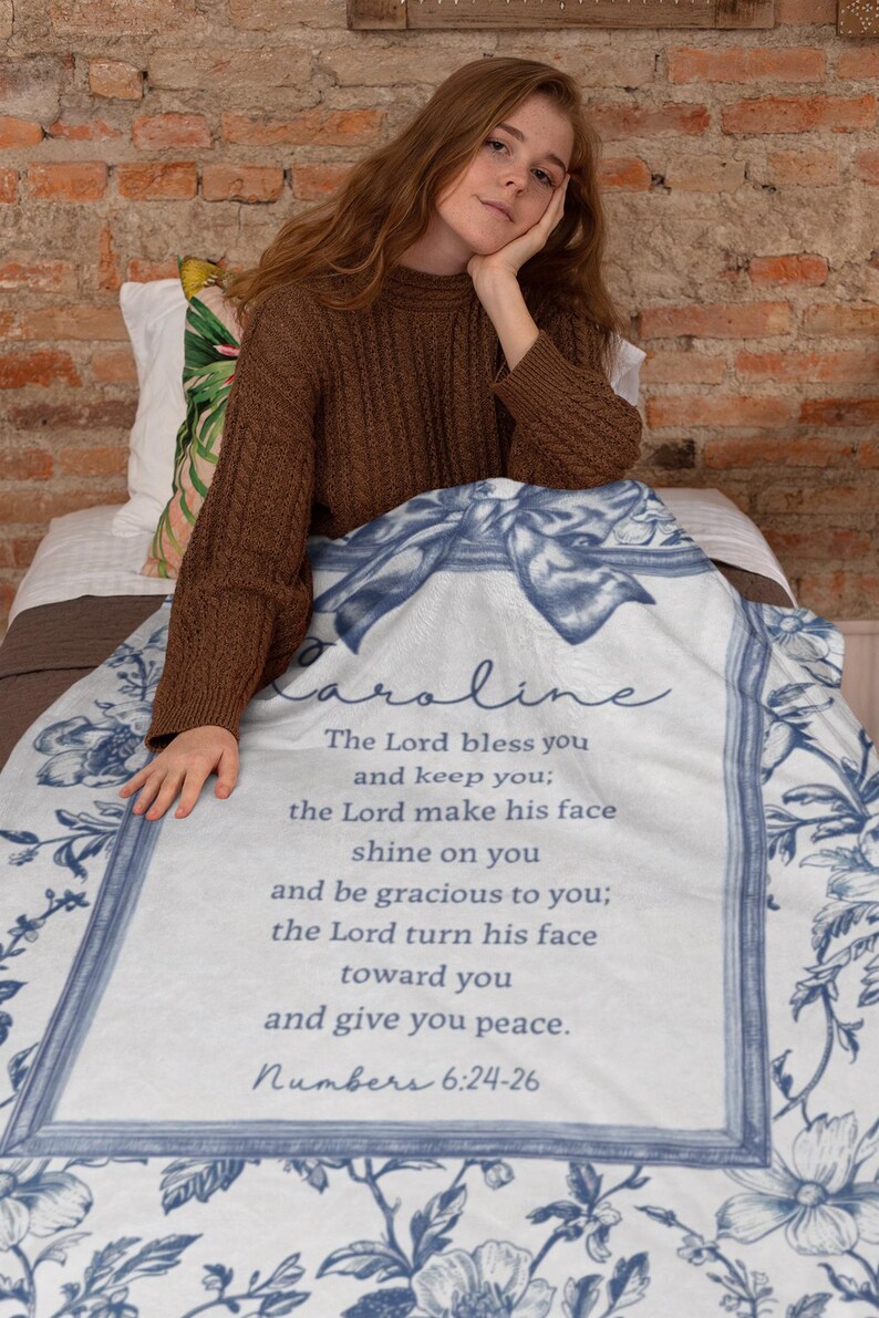 May include: A white and blue floral blanket with the name "Caroline" and a Bible verse printed on it. The verse reads "The Lord bless you and keep you... Numbers 6:24-26". The blanket has a decorative border and bow design.