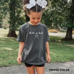 May include: A dark gray t-shirt with the text "Child of God" in white script and a bow graphic. The shirt is a pepper color and has short sleeves. The image shows a young person wearing the shirt.