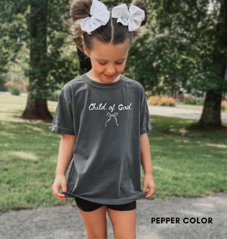 May include: A dark gray t-shirt with the text "Child of God" in white script and a bow graphic. The shirt is a pepper color and has short sleeves. The image shows a young person wearing the shirt.