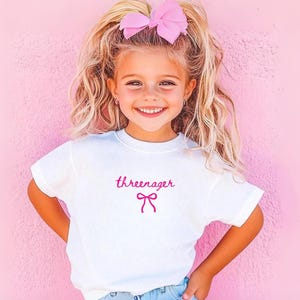 May include: A white t-shirt with the word "threenager" in pink script and a pink bow graphic. The child is wearing a large pink bow in her hair and smiling. The background is a solid pink wall.