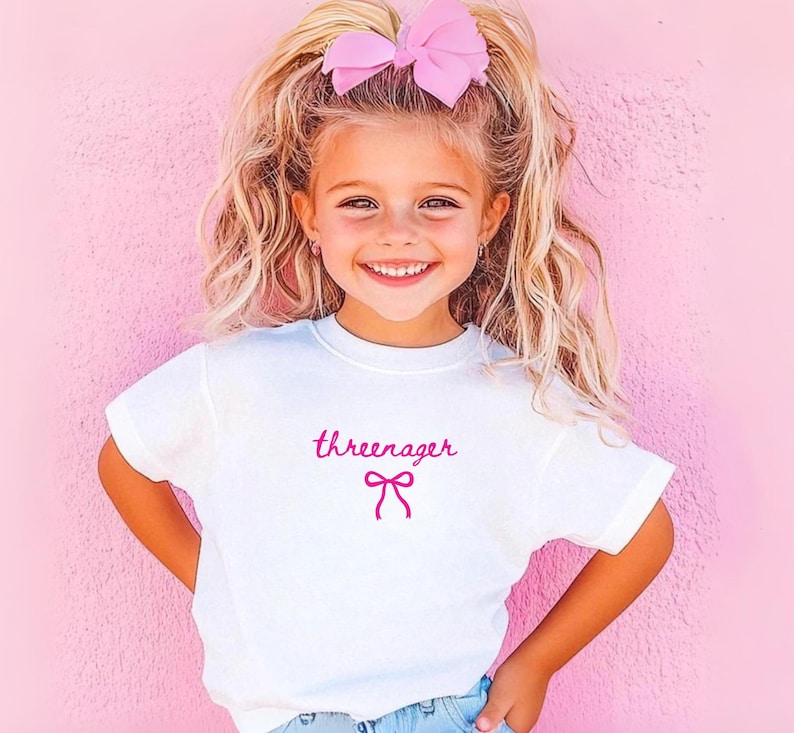 May include: A white t-shirt with the word "threenager" in pink script and a pink bow graphic. The child is wearing a large pink bow in her hair and smiling. The background is a solid pink wall.