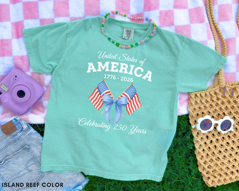 May include: A light green t-shirt with the text "United States of AMERICA 1776-2026 Celebrating 250 Years" and two American flags. The shirt is on a pink and white checkered blanket with a purple camera, denim shorts, and a straw bag.
