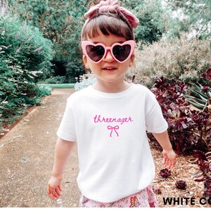 May include: A white t-shirt with the word "threenager" in pink script and a pink bow graphic. The child is wearing pink heart-shaped sunglasses and a pink headband. The background includes greenery and a pathway.