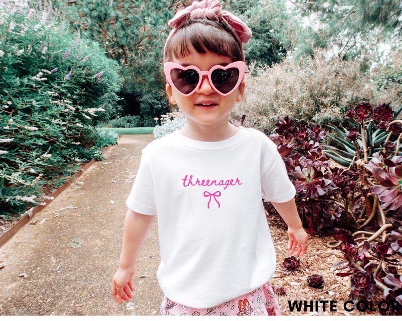 May include: A white t-shirt with the word "threenager" in pink script and a pink bow graphic. The child is wearing pink heart-shaped sunglasses and a pink headband. The background includes greenery and a pathway.