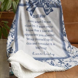 Pode incluir: Um cobertor floral branco e azul com o texto "DEAR MOM" e uma mensagem sincera. O cobertor tem uma parte inferior macia e fofa e est&aacute; sobre um sof&aacute; de couro marrom. O design inclui um la&ccedil;o e uma borda floral.