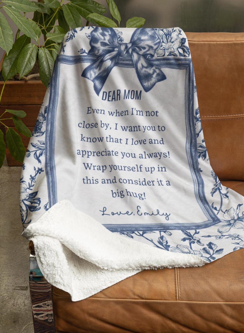 Pode incluir: Um cobertor floral branco e azul com o texto "DEAR MOM" e uma mensagem sincera. O cobertor tem uma parte inferior macia e fofa e est&aacute; sobre um sof&aacute; de couro marrom. O design inclui um la&ccedil;o e uma borda floral.