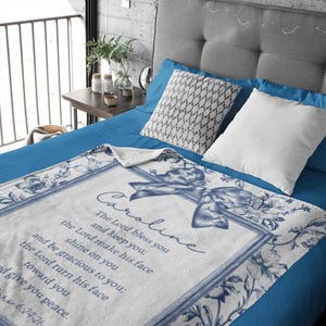 May include: White and blue floral throw blanket with the name "Caroline" and a Bible verse printed on it. The blanket has a blue border and a bow design. The blanket is on a blue bed.