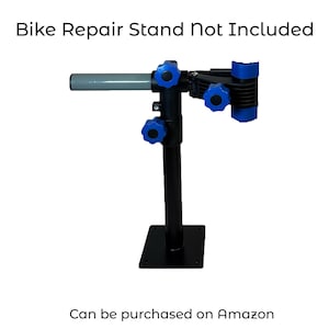 Pode incluir: Um suporte de reparo de bicicleta preto com bot&otilde;es de ajuste azuis e uma al&ccedil;a cinza. O suporte &eacute; montado em uma base quadrada preta. O texto na parte superior diz "Bike Repair Stand Not Included". O texto na parte inferior diz "Can be purchased on Amazon".