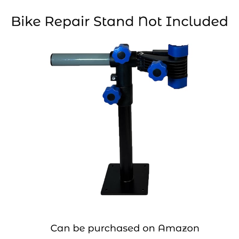 Pode incluir: Um suporte de reparo de bicicleta preto com bot&otilde;es de ajuste azuis e uma al&ccedil;a cinza. O suporte &eacute; montado em uma base quadrada preta. O texto na parte superior diz "Bike Repair Stand Not Included". O texto na parte inferior diz "Can be purchased on Amazon".