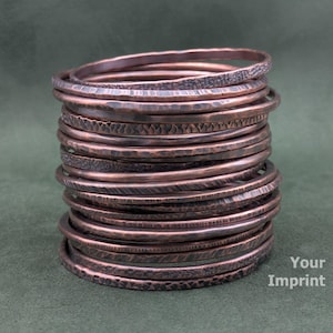 May include: A stack of copper-colored bangles with various textured surfaces. The bangles are stacked on top of each other, showcasing different patterns and finishes. The background is a solid, muted green.