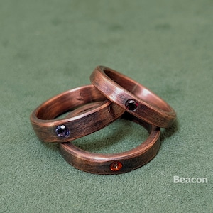 Puede incluir: Tres anillos de cobre con un acabado cepillado y texturizado. Cada anillo presenta una pequeña piedra preciosa redonda y facetada engastada en la banda. Las piedras son de color morado, rojo oscuro y naranja. La palabra Beacon aparece abajo a la derecha.