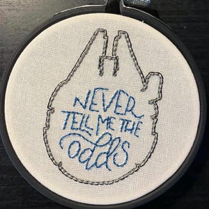May include: Embroidered hoop art with a Millennium Falcon design and the text "NEVER TELL ME THE ODDS" in blue thread. The design is on white fabric, framed by a black hoop.