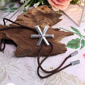 Könnte beinhalten: Ein silberfarbenes Bolo-Tie mit einem sechszackigen Stern-Design. Die Krawatte hat eine braune Kordel und rechteckige Metallspitzen. Das Bolo-Tie wird auf einem Stück Treibholz präsentiert.