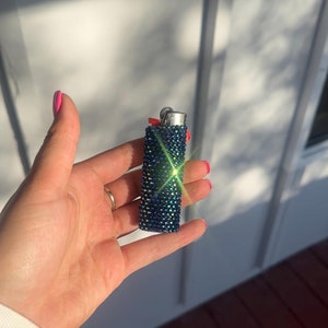 Puede incluir: Un encendedor azul y verde adornado con pedrer&iacute;a. El encendedor est&aacute; cubierto de peque&ntilde;os y brillantes pedrer&iacute;a. La parte superior del encendedor es plateada y el encendedor se sostiene en una mano con esmalte de u&ntilde;as rosa.