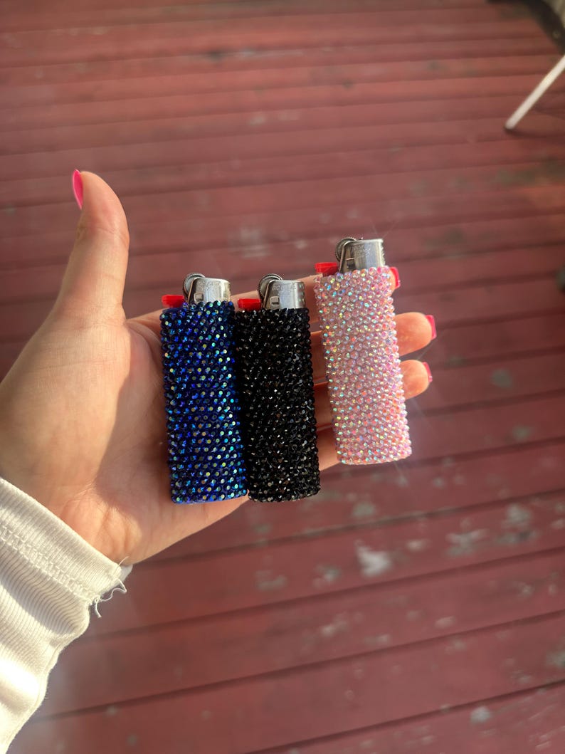 Puede incluir: Tres encendedores enjoyados en azul, negro y rosa. Cada encendedor est&aacute; cubierto de peque&ntilde;os y brillantes pedrer&iacute;a. Los encendedores se sostienen en una mano sobre un fondo de tablones de madera rojos.