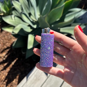 Puede incluir: Un encendedor morado cubierto de pedrer&iacute;a iridiscente. El encendedor tiene una parte superior de metal plateado y un bot&oacute;n de encendido rojo. El fondo presenta plantas verdes y una superficie de madera.