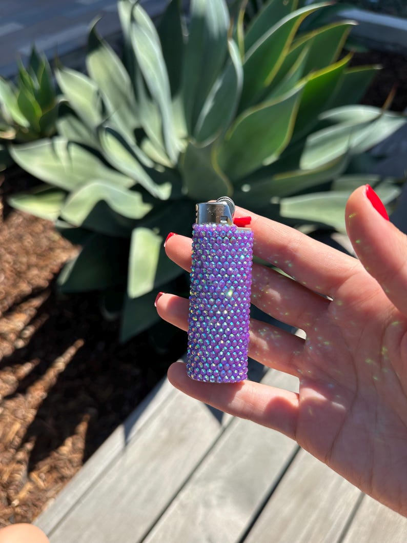 Puede incluir: Un encendedor morado cubierto de pedrer&iacute;a iridiscente. El encendedor tiene una parte superior de metal plateado y un bot&oacute;n de encendido rojo. El fondo presenta plantas verdes y una superficie de madera.