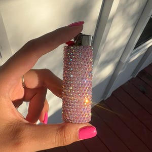 Puede incluir: Un encendedor rosa cubierto de pedrer&iacute;a iridiscente. El encendedor se sostiene en una mano con esmalte de u&ntilde;as rosa. El fondo incluye una pared blanca y una terraza de madera.