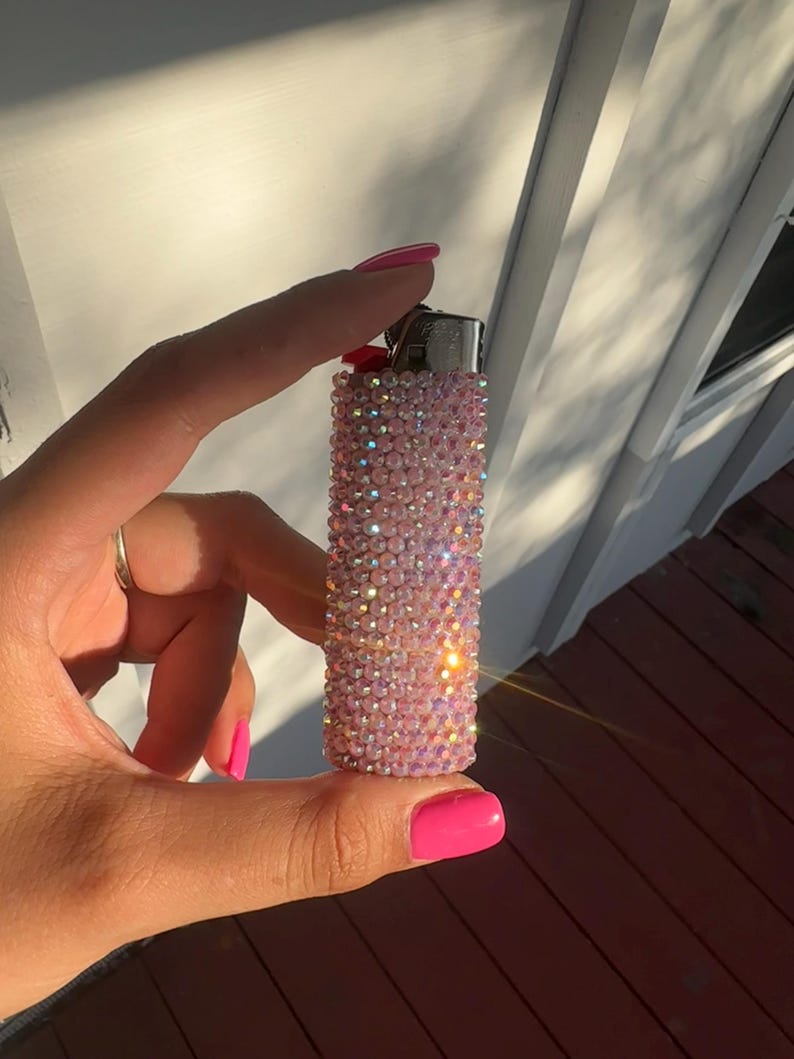 Puede incluir: Un encendedor rosa cubierto de pedrer&iacute;a iridiscente. El encendedor se sostiene en una mano con esmalte de u&ntilde;as rosa. El fondo incluye una pared blanca y una terraza de madera.