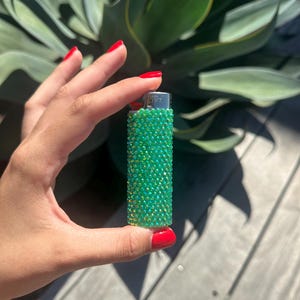 Puede incluir: Un encendedor verde enjoyado. El encendedor est&aacute; cubierto de peque&ntilde;os pedrer&iacute;a verde iridiscente. El encendedor se sostiene en una mano con esmalte de u&ntilde;as rojo, con una planta verde en el fondo.