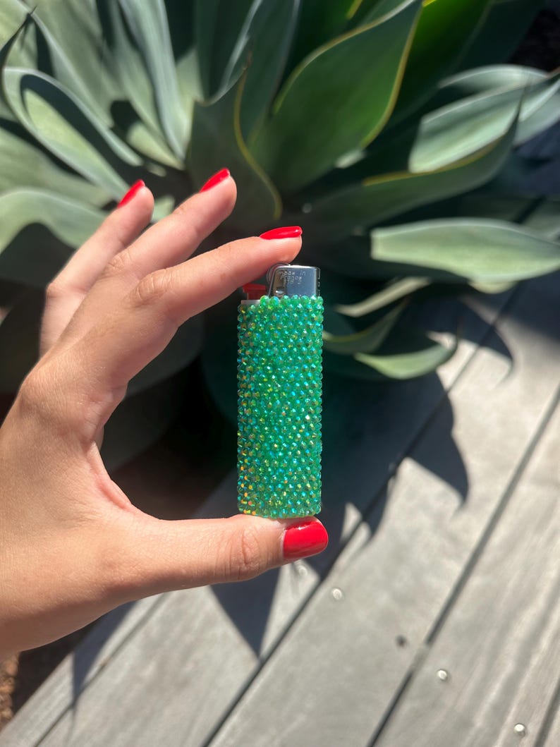Puede incluir: Un encendedor verde enjoyado. El encendedor est&aacute; cubierto de peque&ntilde;os pedrer&iacute;a verde iridiscente. El encendedor se sostiene en una mano con esmalte de u&ntilde;as rojo, con una planta verde en el fondo.