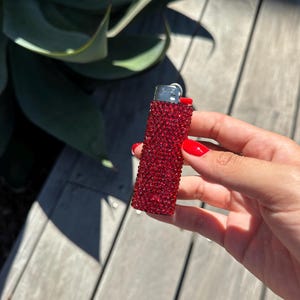 Puede incluir: Un encendedor rojo adornado con pedrer&iacute;a. El encendedor est&aacute; cubierto de peque&ntilde;os pedrer&iacute;a rojos brillantes. La parte superior del encendedor es plateada. El fondo incluye hojas verdes y una superficie de madera.