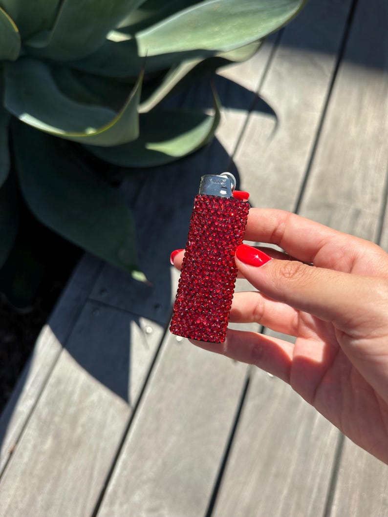 Puede incluir: Un encendedor rojo adornado con pedrer&iacute;a. El encendedor est&aacute; cubierto de peque&ntilde;os pedrer&iacute;a rojos brillantes. La parte superior del encendedor es plateada. El fondo incluye hojas verdes y una superficie de madera.