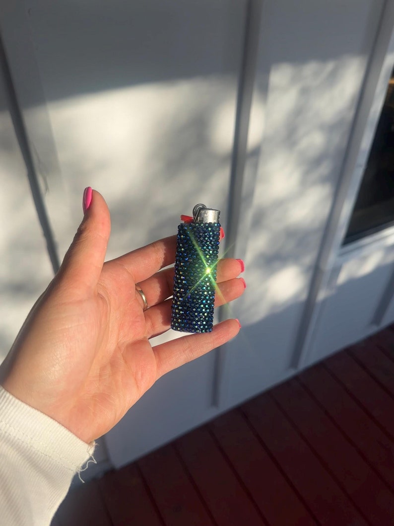 Puede incluir: Un encendedor azul y verde adornado con pedrer&iacute;a. El encendedor est&aacute; cubierto de peque&ntilde;os y brillantes pedrer&iacute;a. La parte superior del encendedor es plateada y el encendedor se sostiene en una mano con esmalte de u&ntilde;as rosa.
