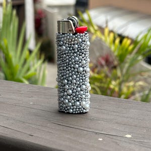 Puede incluir: Un encendedor plateado cubierto con perlas de imitaci&oacute;n de varios tama&ntilde;os. La parte superior del encendedor tiene una carcasa de metal plateado con un bot&oacute;n de encendido rojo. El encendedor est&aacute; de pie sobre una superficie de madera.
